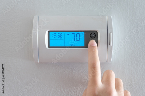 A woman is pressing the down button of a wall attached house thermostat with digital display showing temperature 70 degree Fahrenheit for heating, cooling, electricity and gas saving photo