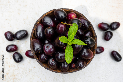 Dark pink-red ripe Syzygium cumini fruits. Dark black java plum in a wood bowl at isolated white background. Green mint leaf on top of some large java plums. photo