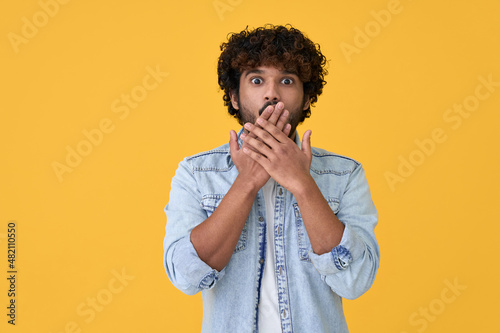 Shocked surprised young indian man looking at camera with omg face expression covering mouth with hands feeling amazed speachless silent reaction standing isolated on yellow background. photo