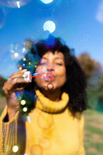 Vertical portrait of young african american woman making soap bubbles outdoors. Selective focus on bubbles.