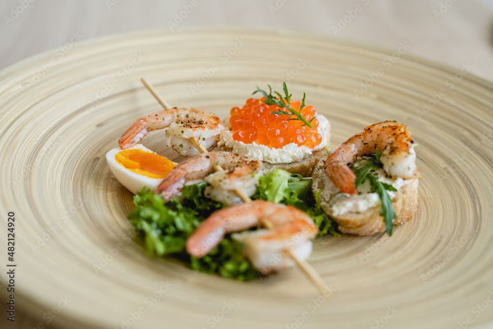 A delicious homemade grilled shrimps on a skewer, toasts with curd cheese, King prawn, red caviar and arugula. Plate with seafood, sliced eggs, green fresh lettuce, arugula and toasts