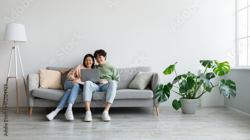 Loving Asian couple sitting on couch, using laptop together, looking at computer screen, watching movie on web at home