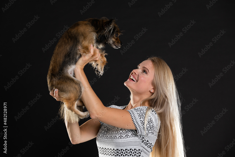 A smiling blonde woman lifts her dog up and they look into each other's eyes. Long hair. Black background.