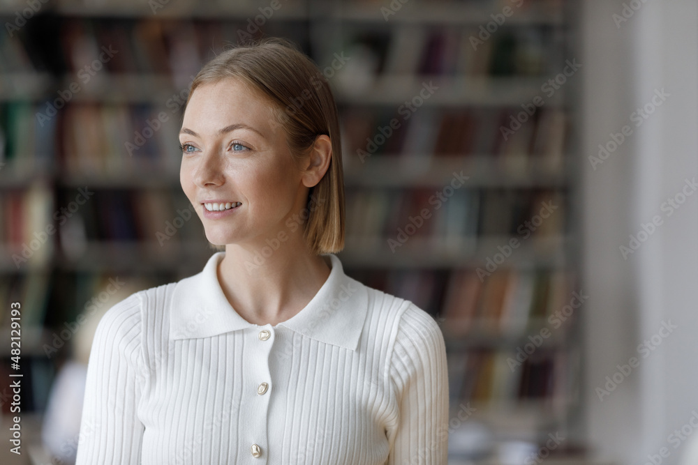 Happy thoughtful millennial university student girl standing in public library, looking away, smiling at good thoughts, thinking over research study project, future college degree Happy thoughtful millennial university student girl standing in public library, looking away, smiling at good thoughts, thinking over research study project, future college degree