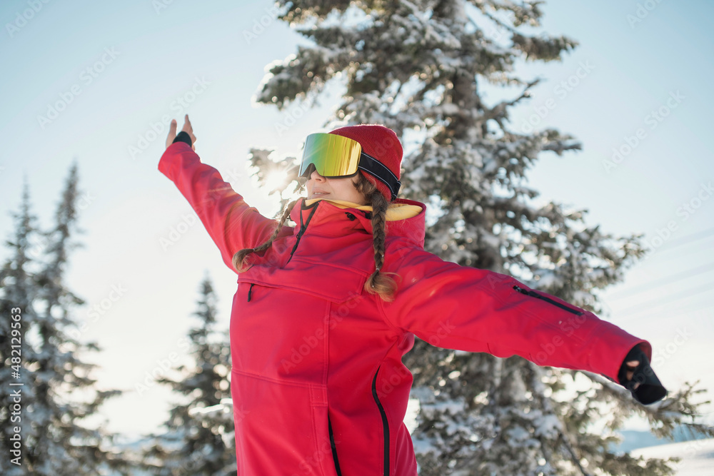A woman wearing ski outfit between pine trees. Winter and snow. Ski resort concept. Woman wearing ski mask.