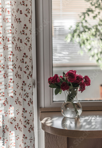 A bouquet of garden roses in a vase with water on a window sill against the background of a window and a loggia with a Benjamin ficus  a curtain in a small flower.