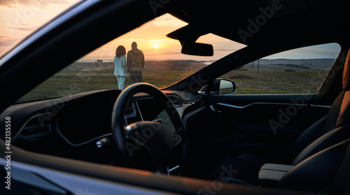 Back view of young couple in love enjoying sunset automobile date at nature environment, husband and wife spending evening for bonding togetherness with black car for trip travelling on front © anatoliy_gleb
