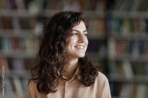 Happy dreamy beautiful student looking away, smiling at good thoughts, thinking over future university degree. Thoughtful Hispanic college girl visiting library head shot portrait