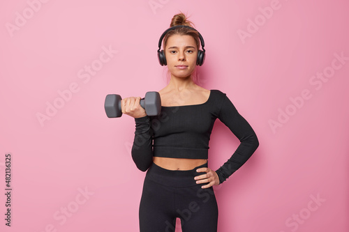 Horizontal shot of motivated sporty woman dressed in black top and legings raises dumbbell indoor trains muscles listens music via headphones isolated over pink background does sport exercises