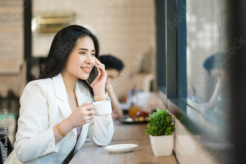 Asian businesswoman using technology. Professional woman is conversing on her phone while drinking coffee in a coffee shop. Working from anywhere