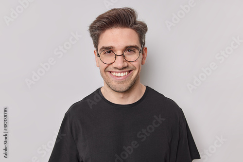 Portrait of happy man with dark hair smiles with white teeth looks confident at camera wears round transparent glasses black t shirt poses at studio against white background. Positive emotions © wayhome.studio 