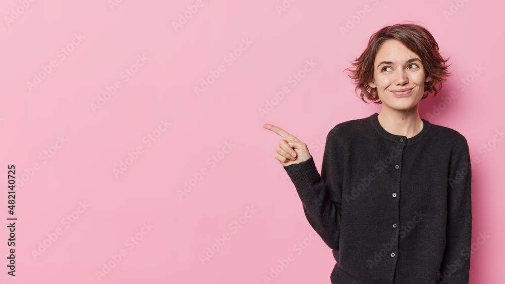 Horizontal shot of positive young European woman points away at blank space on left asks out for coffee invites check out something dressed in casual black jumper isolated over pink backgound Horizontal shot of positive young European woman points away at blank space on left asks out for coffee invites check out something dressed in casual black jumper isolated over pink backgound