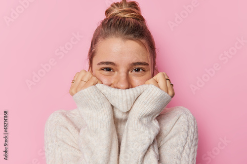 Positive young European woman with fair hair gathered in bun wears knitted warm white sweater during cold temperature weather covers mouth with collar looks happily isolated over pink background