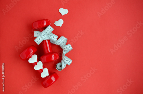 Two red dumbbells, white measuring tape, hearts on red background with copy space. Concept of Valentines day, healthy lifestyle, giving gifts, love of sports, shopping