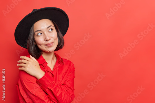 Thoughtful dreamy Asian woman with dark hair wears black hat and shirt keeps hand on shoulder looks away pensively considers something isolated over vivid red background with blank copy space