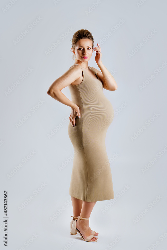 Fototapeta premium Studio portrait of an elegant young adult pregnant woman in a beige dress on a gray background, the concept of a happy pregnancy.