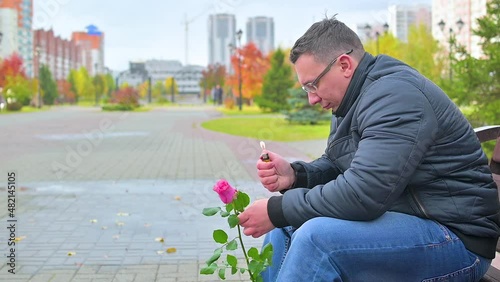 A man with a rose sits on a bench and looks at the fire in a lighter in the pouring rain in autumn