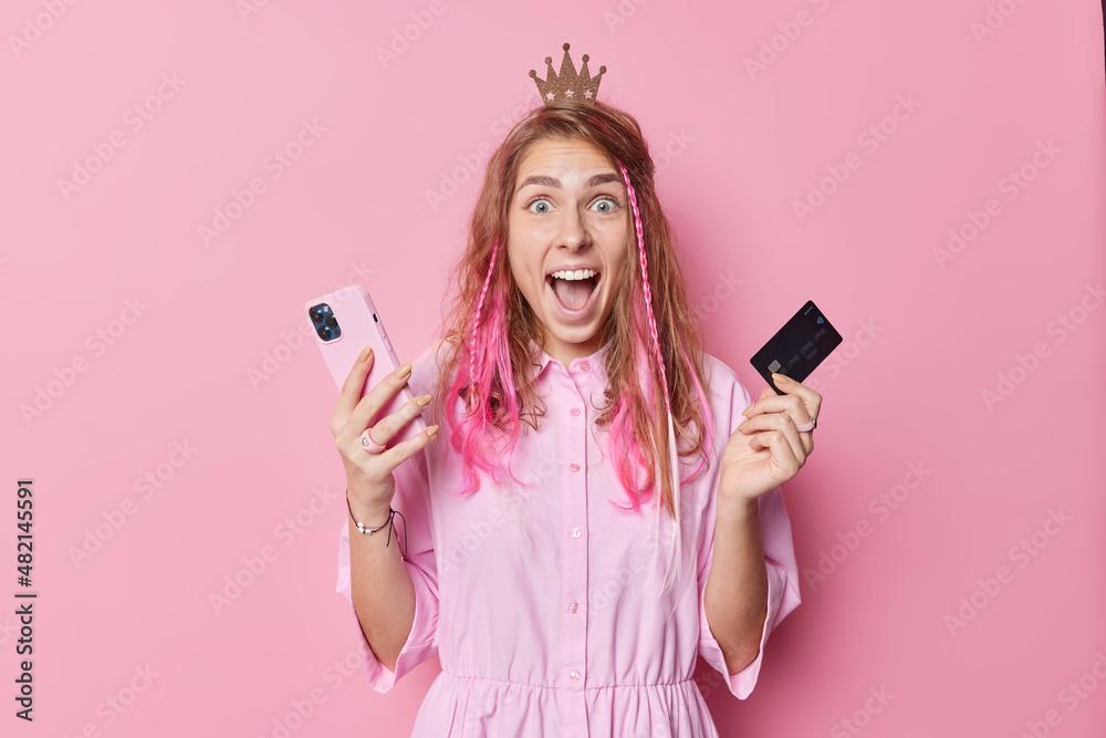 Surprised cheerful woman holds mobile phone and credit card makes online shopping glad to receive discount wears crown shirt isolated over pink background gets good order internet wins money online Surprised cheerful woman holds mobile phone and credit card makes online shopping glad to receive discount wears crown shirt isolated over pink background gets good order internet wins money online