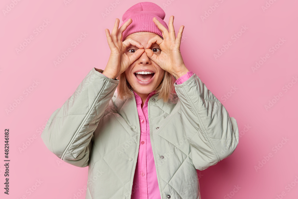 Positive playful cute woman makes goggles with fingers over eyes being childish and optimistic wears hat and jacket isolated over pink background foolishes around and wastes time. Lets have fun Positive playful cute woman makes goggles with fingers over eyes being childish and optimistic wears hat and jacket isolated over pink background foolishes around and wastes time. Lets have fun