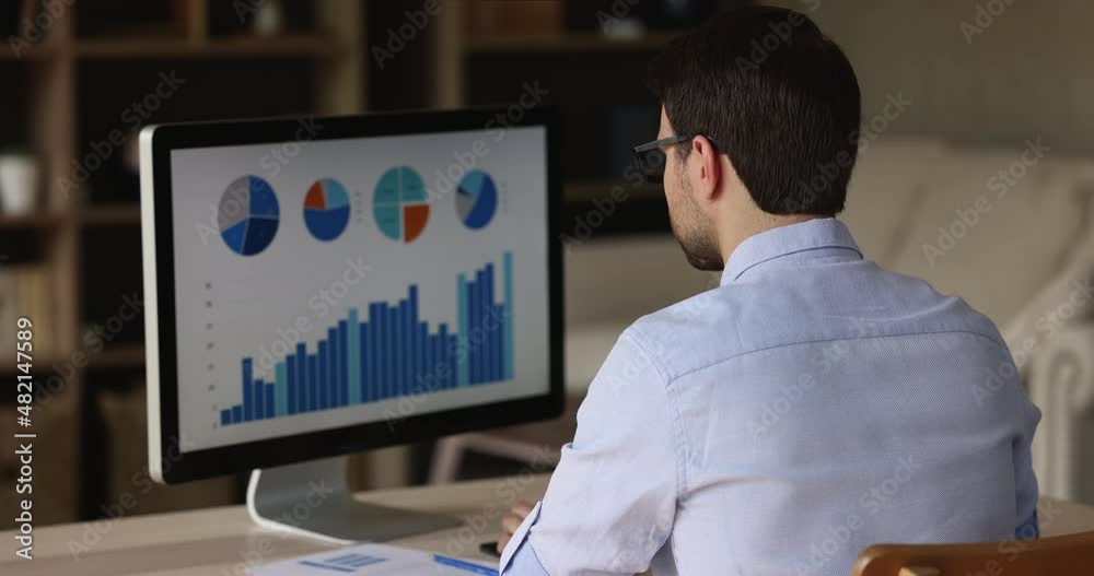 Rear view businessman sit at desk analyze infographic diagrams, seen on pc monitor, working with statistic shown on charts and graphs, computer screen over male shoulder. Sales, company growth concept