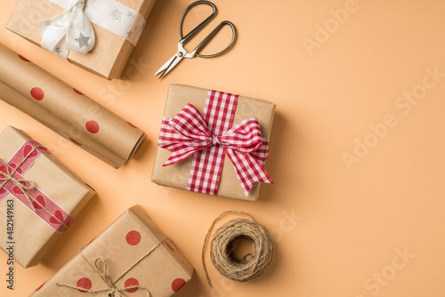 Top view photo of st valentine's day decor craft paper gift boxes with bows scissors spool of twine and roll of kraft paper with polka dot pattern on isolated beige background with copyspace