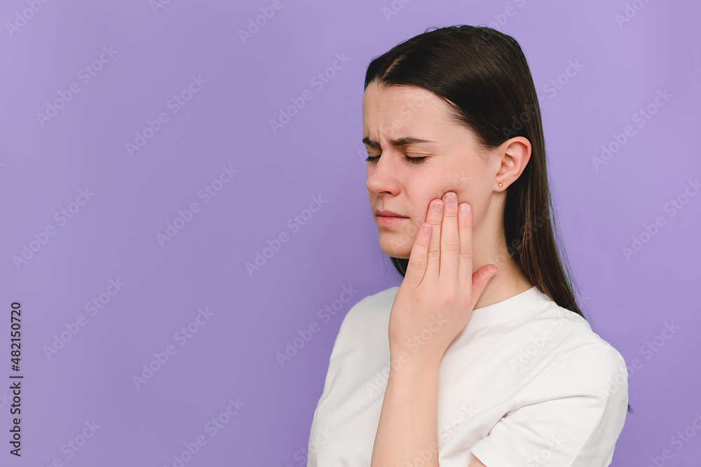 Portrait of unhappy sad young brunette female suffering from acute tooth pain, touching her sore jaw and frowning, model over purple studio background with copy space. Oral hygiene, tooth care concept Portrait of unhappy sad young brunette female suffering from acute tooth pain, touching her sore jaw and frowning, model over purple studio background with copy space. Oral hygiene, tooth care concept
