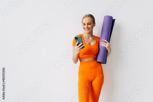 Young athletic woman holding rubber mat for yoga and using mobile phone, standing against white background