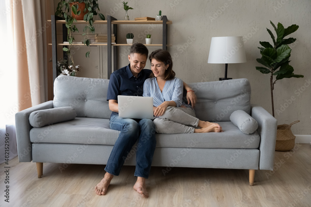 Joyful bonding young family couple using laptop, resting on comfortable couch, enjoying web surfing information, booking flight tickets or hotel online planning trip, shopping in internet store. Joyful bonding young family couple using laptop, resting on comfortable couch, enjoying web surfing information, booking flight tickets or hotel online planning trip, shopping in internet store.