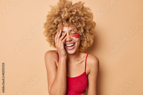 Overjoyed curly haired woman covers face with palm smiles broadly applies beauty patches under eyes for skin rejuvenation dressed in top isolated over beige background undergoes facial treatments