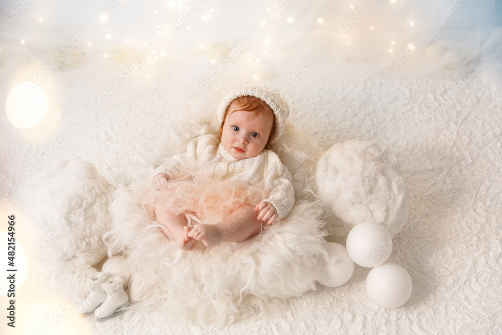 Little red hair baby on the white wool. Infant girl wearing white sweater and hat in photo studio which makes like winter composition. Garlands are behind the child. Little red hair baby on the white wool. Infant girl wearing white sweater and hat in photo studio which makes like winter composition. Garlands are behind the child.