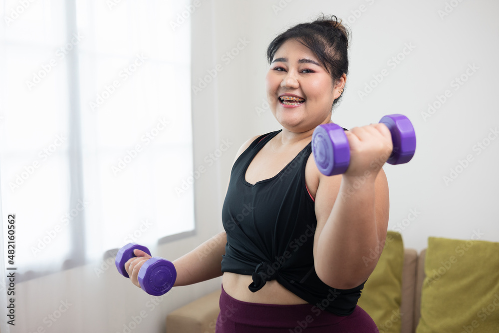 Attractive asian young fitness woman plus size lifting dumbbell weights workout at home in living room. Female training and exercise wearing sport wear fit body. Happy and is proud of herself. Attractive asian young fitness woman plus size lifting dumbbell weights workout at home in living room. Female training and exercise wearing sport wear fit body. Happy and is proud of herself.