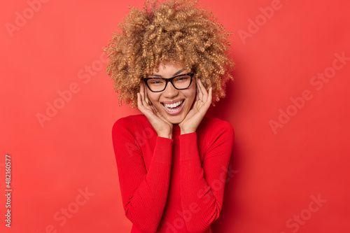 Happy positive young curly haired woman smiles broadly keeps hands on cheeks being in good mood wearrs transparent glasses and jumper isolated over vivid red background. Happy emotions concept