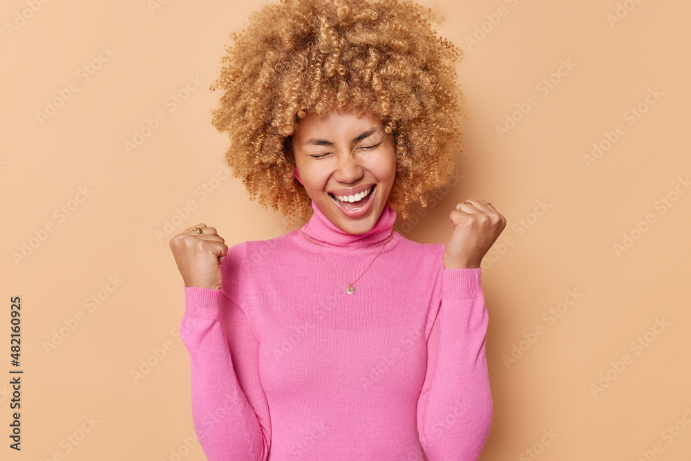 Glad curly haired woman clenches fists with triumpg feels like winner celebrates triumph wears casual pink turtleneck isolated over beige background rejoices victory achieves goals. Yes I did it