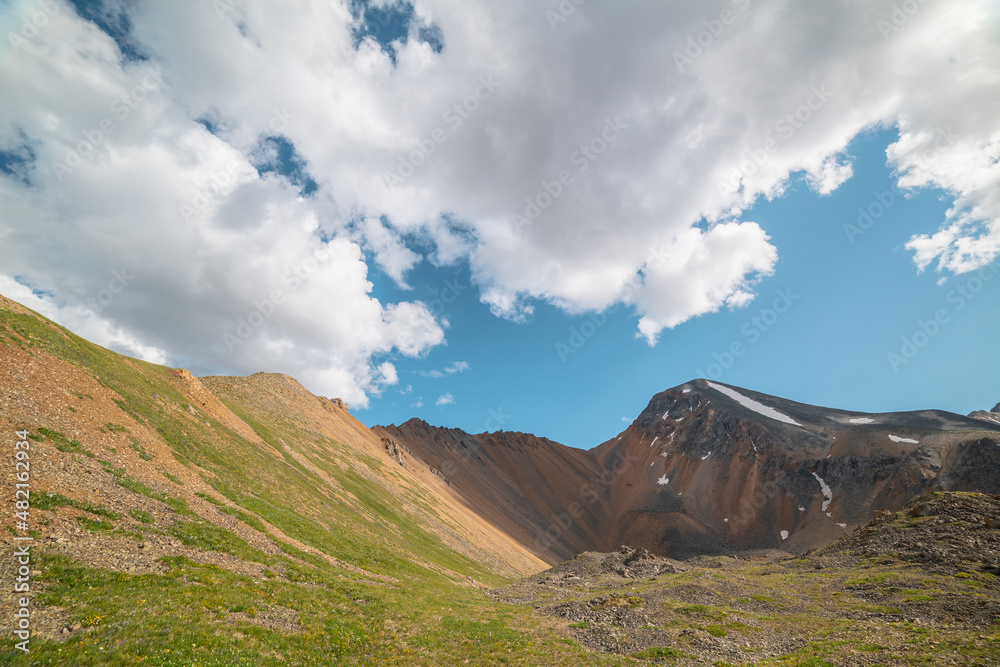 Scenic landscape with sunlit green grassy stone hill and high orange mountain ridge under white clouds in blue sky. Colorful scenery with rocky mountains and sharp rocks in sunlight under cloudy sky. Scenic landscape with sunlit green grassy stone hill and high orange mountain ridge under white clouds in blue sky. Colorful scenery with rocky mountains and sharp rocks in sunlight under cloudy sky.