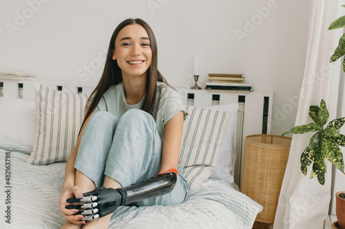 Laughing gorgeous European young lady sitting on bed with legs in her hands, having bionic prosthetic arm, long beautiful brown hair, wearing casual clothes, resting at home in bedroom