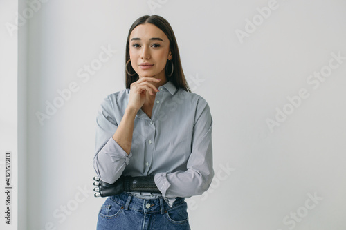 Horizontal portrait of shy gorgeous young lady in blue jeans and shirt holding chin, posing with bionic prosthetic arm, standing against white studio wall with copy space for your advertising content