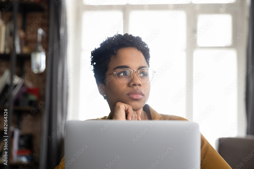 Pensive millennial African employee woman in glasses distracting from work at laptop, looking away with thoughtful face, touching chin, thinking of project future vision, planning business tasks Pensive millennial African employee woman in glasses distracting from work at laptop, looking away with thoughtful face, touching chin, thinking of project future vision, planning business tasks