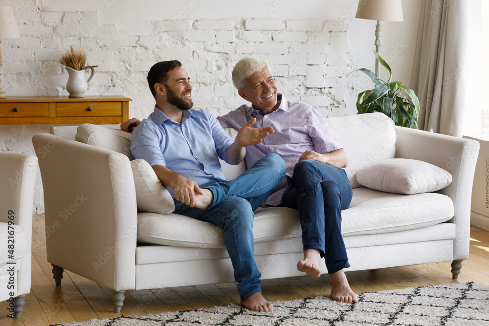 Happy grownup son visiting senior 70s father. Grey haired dad and adult male child relaxing on couch at home, talking, chatting, discussing family plans, events, looking away, laughing Happy grownup son visiting senior 70s father. Grey haired dad and adult male child relaxing on couch at home, talking, chatting, discussing family plans, events, looking away, laughing