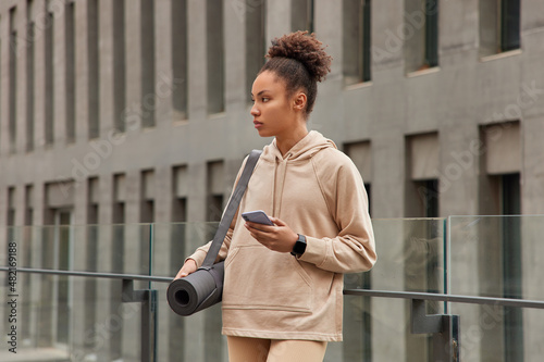 Horizontal shot of active sporty curly woman trains outdoors dressed in sweatshirt uses modern cellphone and fitness mat poses against modernn building checks newsfeed downloads application. photo