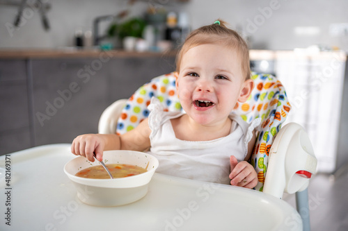 Cute baby girl toddler sitting in the high chair and eating her lunch soup at home kitchen.