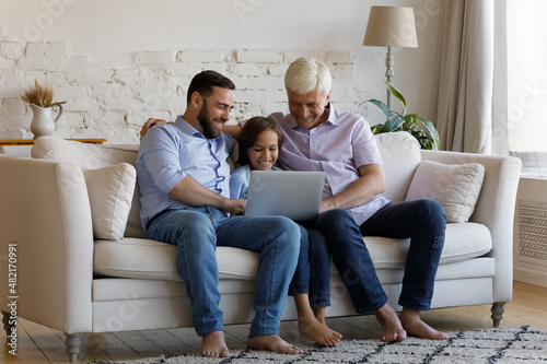 Cheerful grandfather, dad, gen Z kid boy using laptop on couch, relaxing on couch in living room, watching online movie, series, shopping on internet, playing virtual game relation, relationship