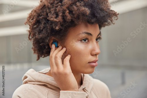 Sideways shot of curly haired thoughtful sportswoman listens music via earphones wears hoodie prepares for training outdoors focused forward poses against blurred background leads sporty lifestyle.