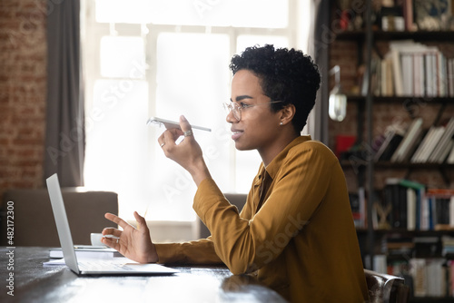 Positive African freelance professional woman having telephone call on speaker, recording audio message on cellphone at table with laptop. Business woman using digital gadgets for work communications