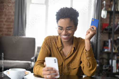 Happy cheerful millennial Black girl enjoying online shopping, using ecommerce app on smartphone, paying for purchase, order by credit card, buying goods on Internet. Finance, spending money concept