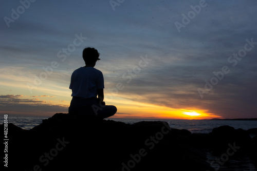 Girl sitting on a rock meditating in the late afternoon near the sea  sun setting in the background. Unrecognizable silhouette. Amazing landscape at dusk. Stones  ocean and sky. Meditation.