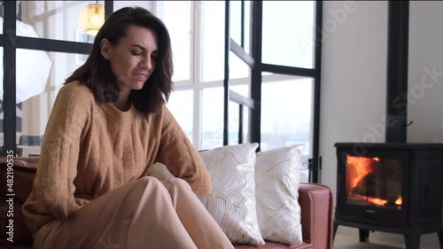 Young woman sitting on the couch at home, suffering from the pain of abdominal cramps, breathing heavily, clutching holding her stomach, face distorted in pain 