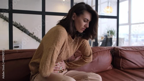 Young woman sitting on the couch at home, suffering from the pain of abdominal cramps, breathing heavily, clutching holding her stomach, face distorted in pain 