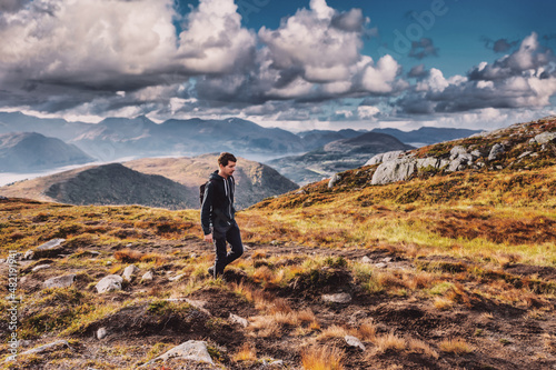 Young adult man hiking on top of a mountain in norway during autumn, wearing a backpack, looking down, sky and horizon with mountain range, volda norway
