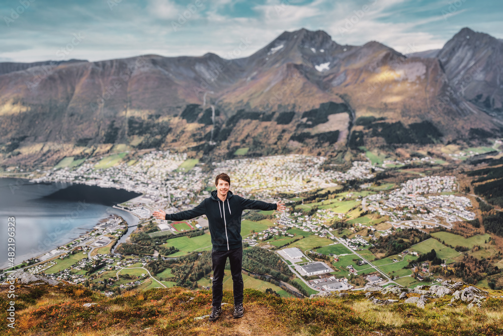 Young adult hiking man standing at the top of a mountain with arms outstretched smiling and looking at camera, mountain range and city in background, norway orsta volda during autumn