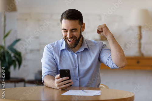 Happy excited smartphone user enjoying win, success, achieve, high job result. Euphoric businessman using cellphone, reading text message, feeling joy, making hand winner gesture, laughing photo